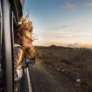beautiful caucasian young woman travel outside the car with wind in the curly hair, motion and movement on the road discovering new places during a nice sunset, enjoy and joyful freedom concept